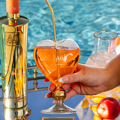 Au Vodka Juicy Peach bottle next to a hand holding a heart shaped glass with orange cocktail and gold straw