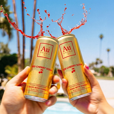 Two Au Vodka Red Cherry Soda cans clinking with red liquid splashing against a blue sky