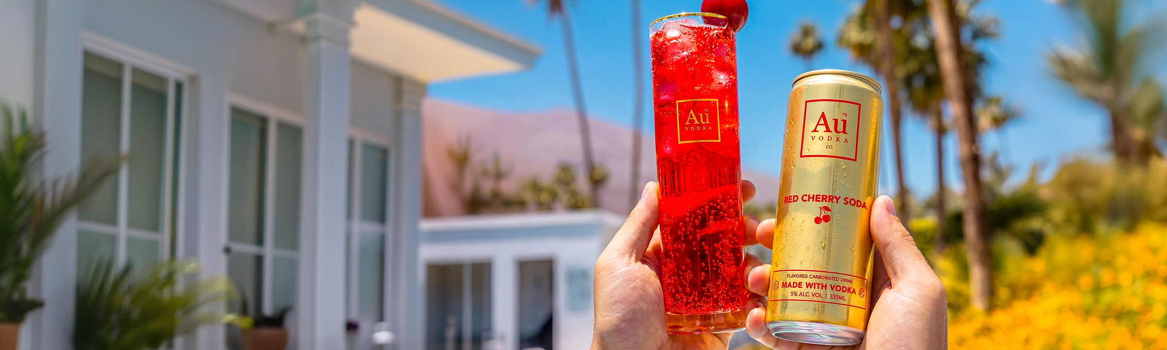 Hands holding a tall glass of red cherry soda and a gold can of Au Vodka Red Cherry Soda