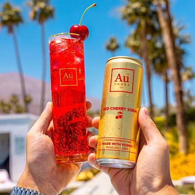 Two hands holding an Au Vodka USA Red Cherry Soda can and a glass with the drink and a cherry
