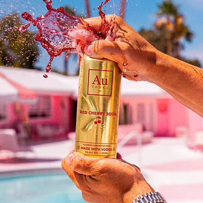 Hands opening an Au Vodka USA Red Cherry Soda can with liquid splashing out, pink buildings in background