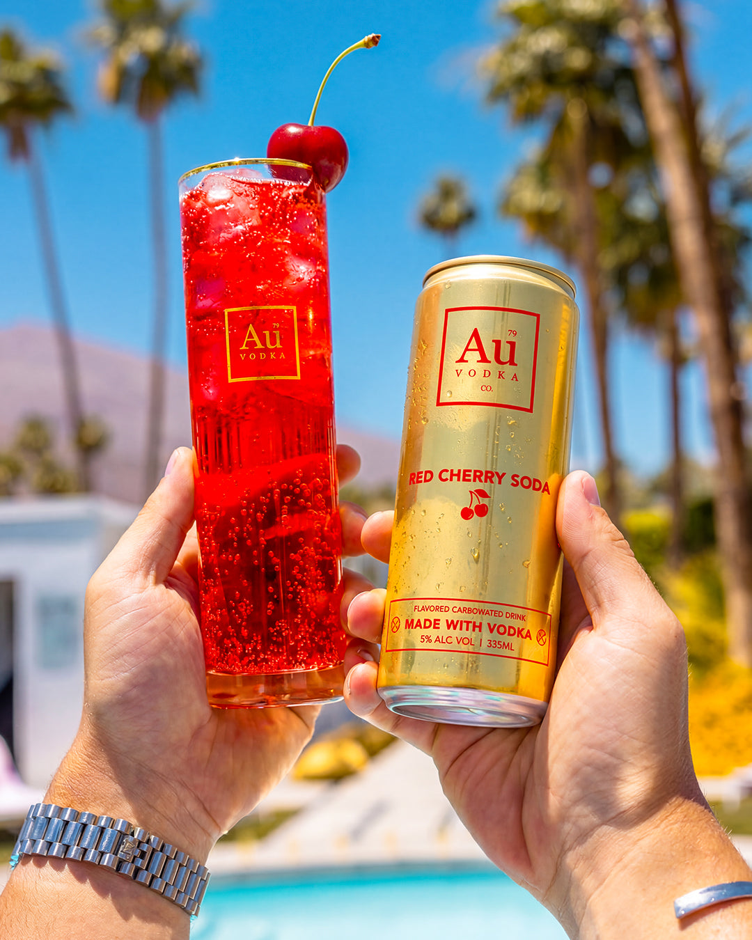 Hands holding a glass of red cherry soda with a cherry garnish and a gold can of Au Vodka Red Cherry Soda