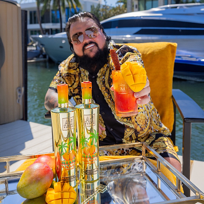 Man in sunglasses holding a mango cocktail, with two Au Vodka Miami Mango bottles and fresh mangoes