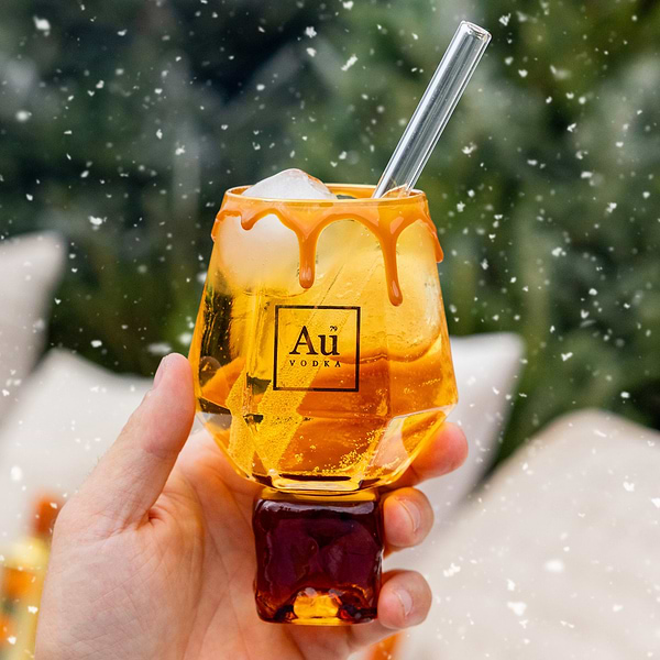 Hand holding a glass of peach flavored liquid with a straw, against a snow and christmas style background.