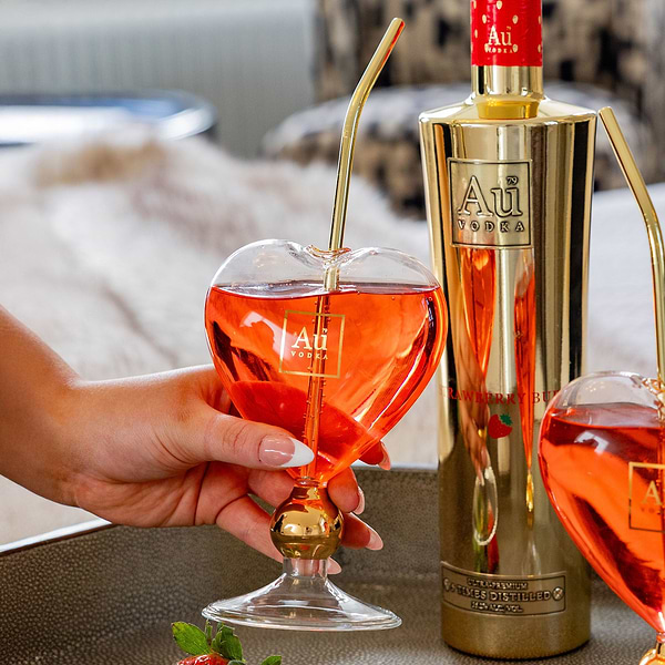 Hand holding heart shaped Au Vodka glass with gold straw next to gold bottle of Au Vodka Strawberry Burst