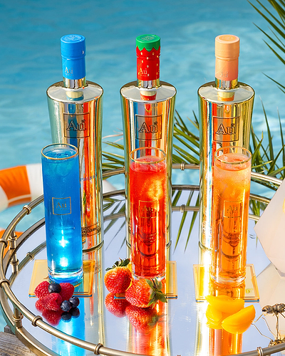 Three Au Vodka bottles and matching cocktails on a mirrored tray by a pool