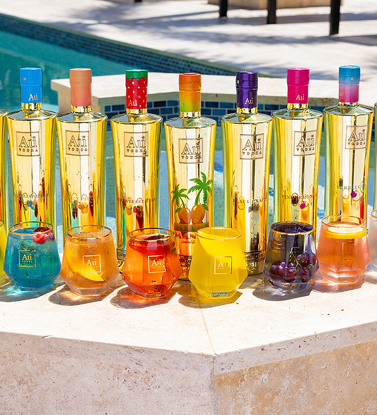 Seven Au Vodka bottles in gold with colorful caps and matching cocktails by a pool