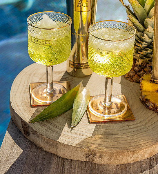 Two Au Vodka drinks with ice in patterned glasses on a wooden table with pineapple and a gold Au Vodka bottle