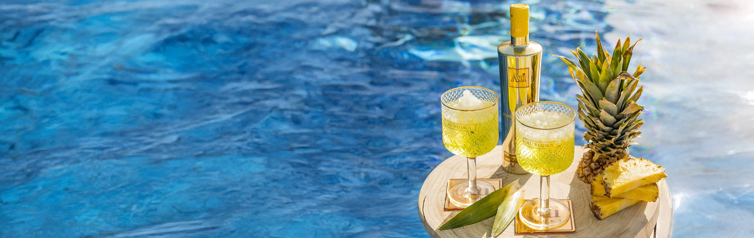 Au Vodka Pineapple Bottle with two drinks and pineapple slices on a wooden table by a pool