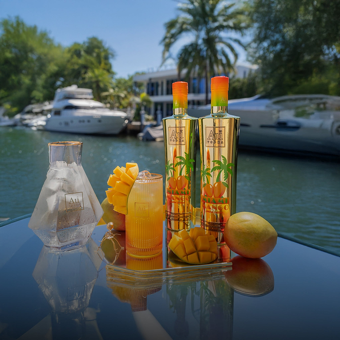 Two Au Vodka Miami Mango bottles, a cocktail, and mangoes on a reflective surface by a canal