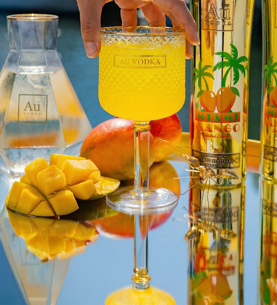 Hand holding a glass of Au Vodka Miami Mango cocktail with mango slices and bottles on a reflective surface