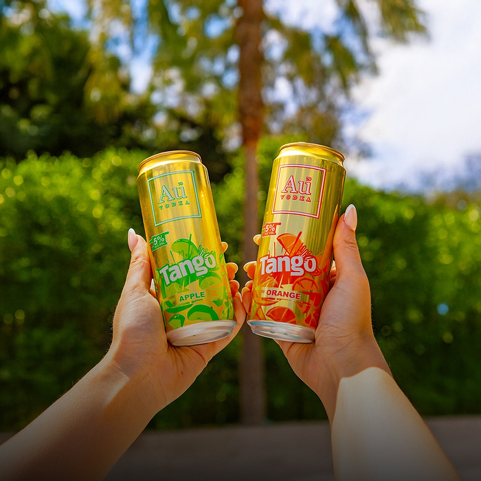 Two hands holding up Au Vodka Tango Apple and Tango Orange cans against a green background