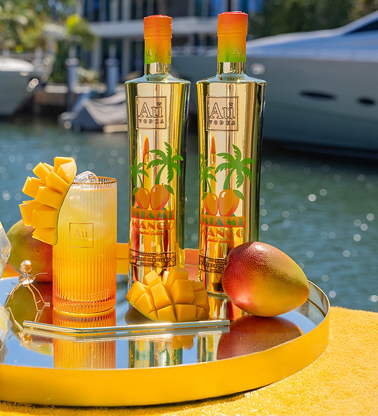 Two Au Vodka Miami Mango bottles, a cocktail, and mango slices on a tray by the water