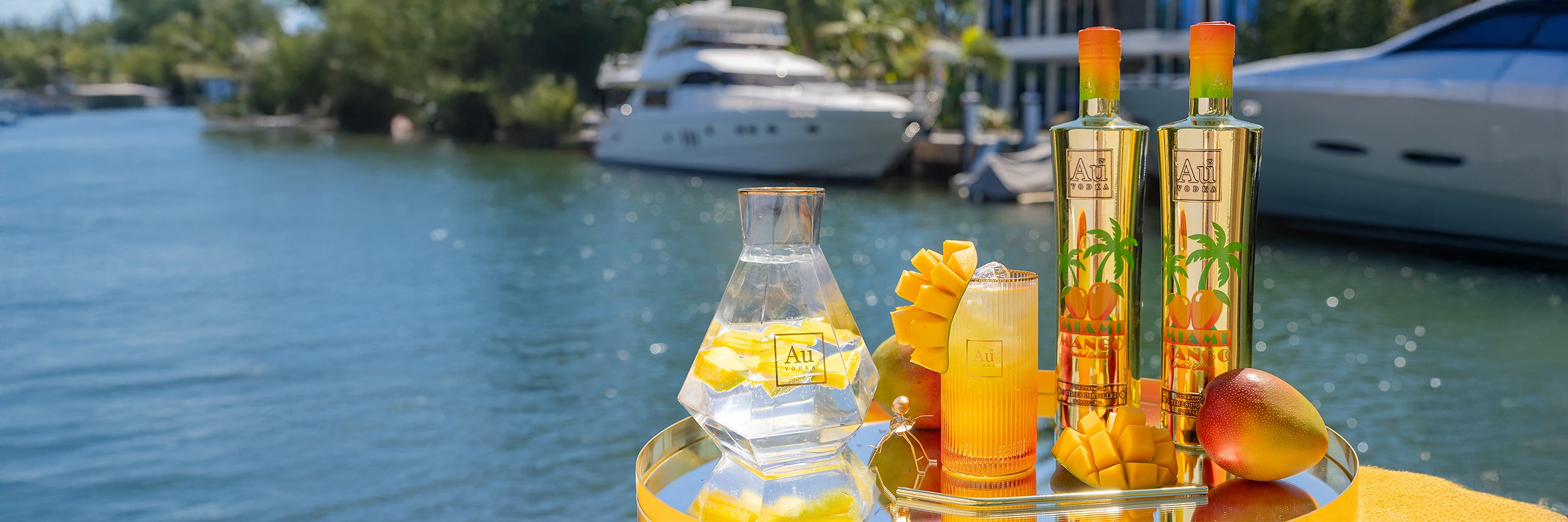 Au Vodka Miami Mango bottles and cocktail on a gold tray by the water with a yacht in the background