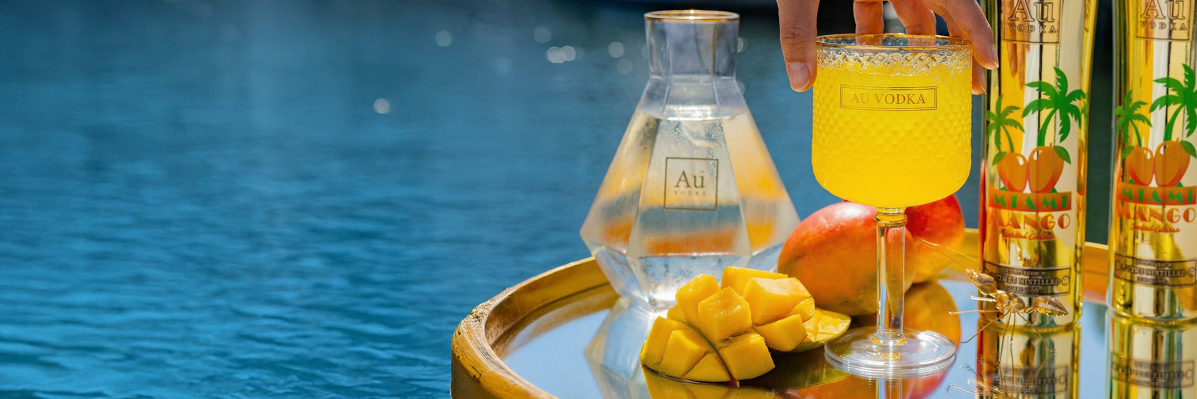 Au Vodka bottle, cocktail, and mangoes on a tray by the water