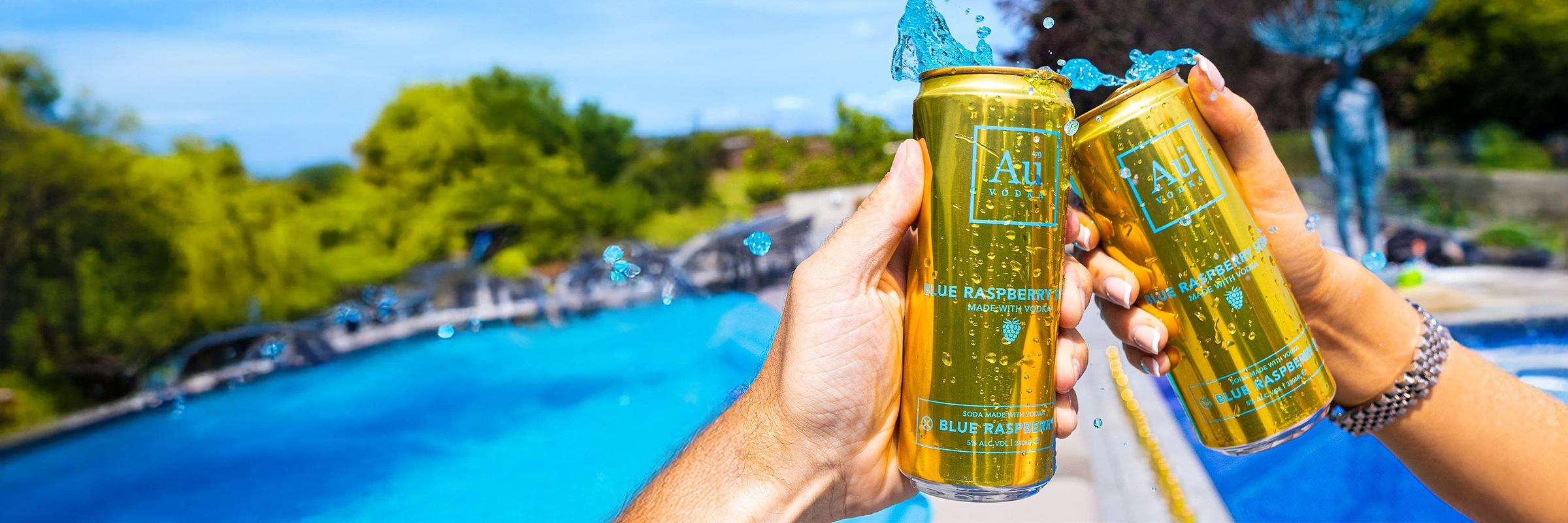 Two hands holding Au Vodka Blue Raspberry cans, splashing blue liquid, by a pool with green trees