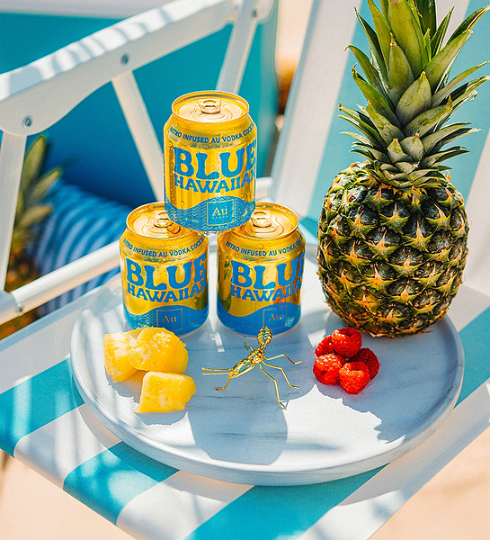 Three Au Vodka Blue Hawaiian cans, a pineapple, pineapple chunks, raspberries, and a gold ant on a tray