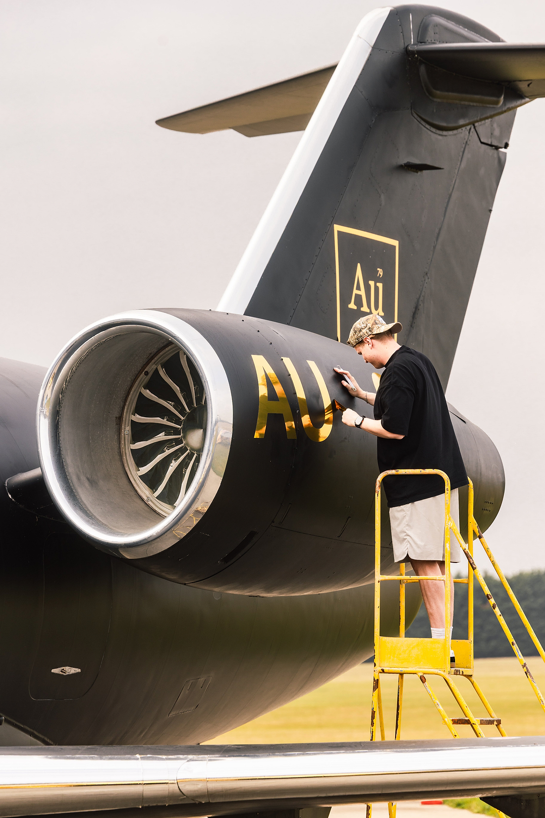 Person standing on a yellow ladder applying gold Au lettering to a black private jet engine