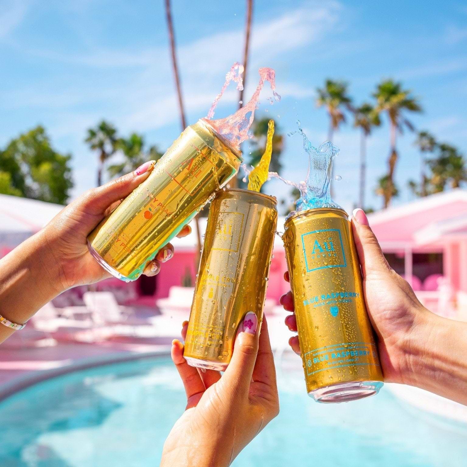 Three hands holding gold Au Vodka cans with splashing liquid against a pool and palm tree background