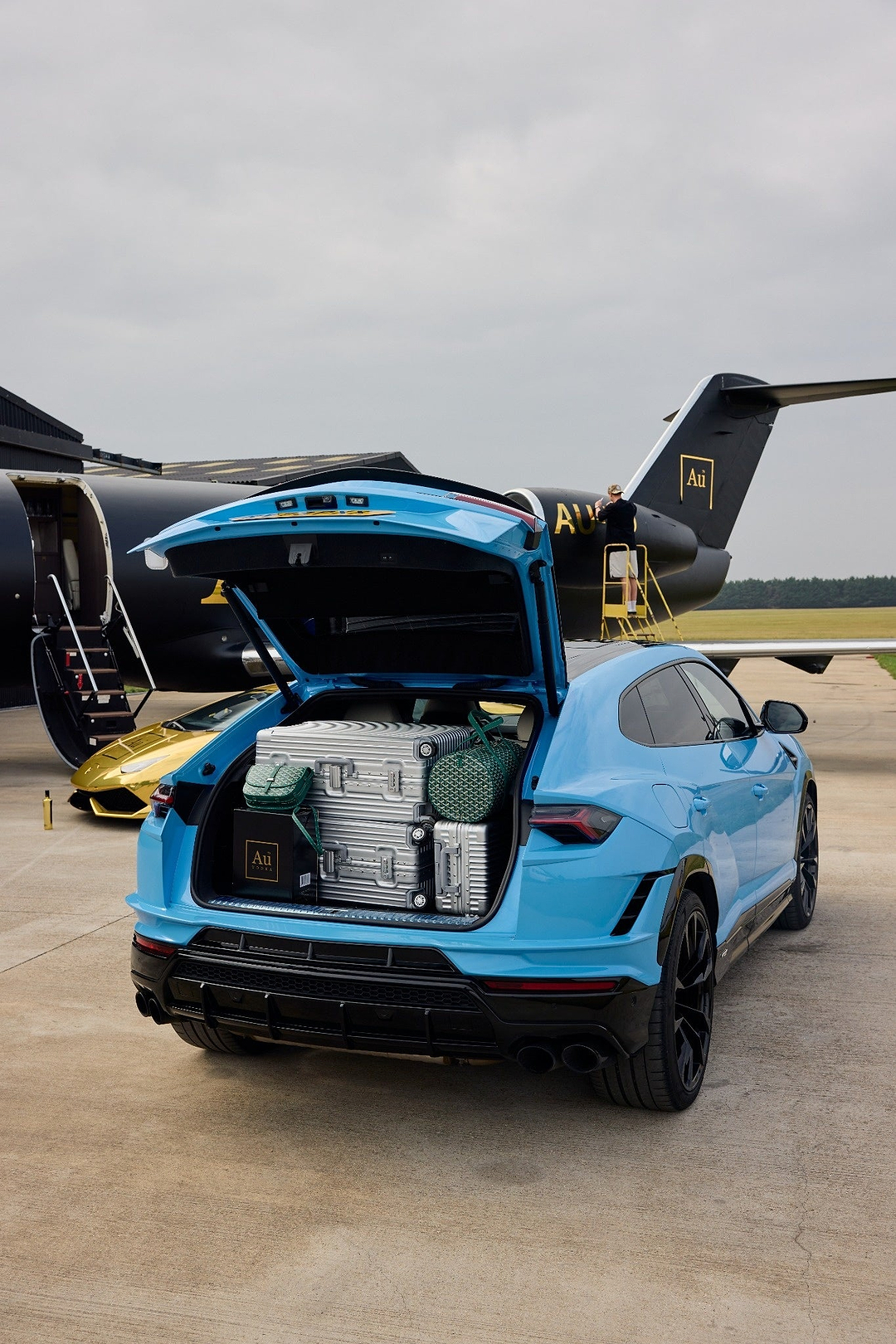 Blue SUV with open trunk packed with silver suitcases and Au Vodka bags parked in front of a private jet