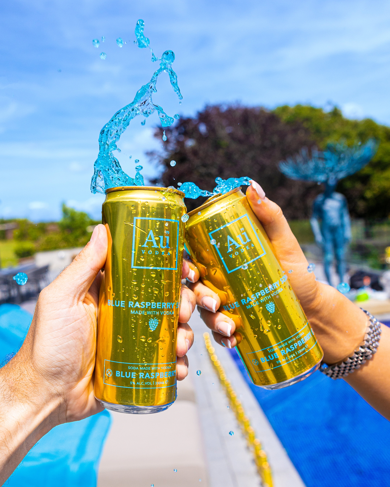 Two hands holding gold Au Vodka Blue Raspberry soda cans with blue liquid splashing outdoors