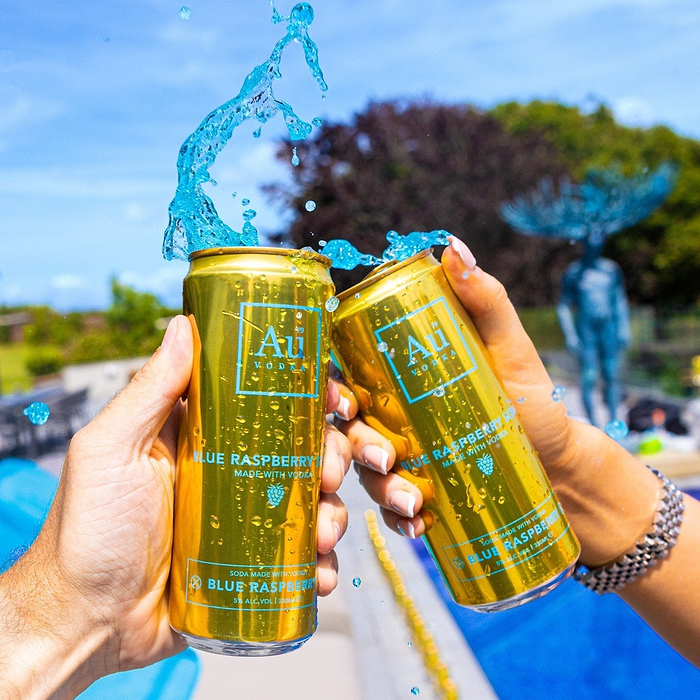 Two hands holding gold Au Vodka Blue Raspberry cocktail cans with blue liquid splashing out
