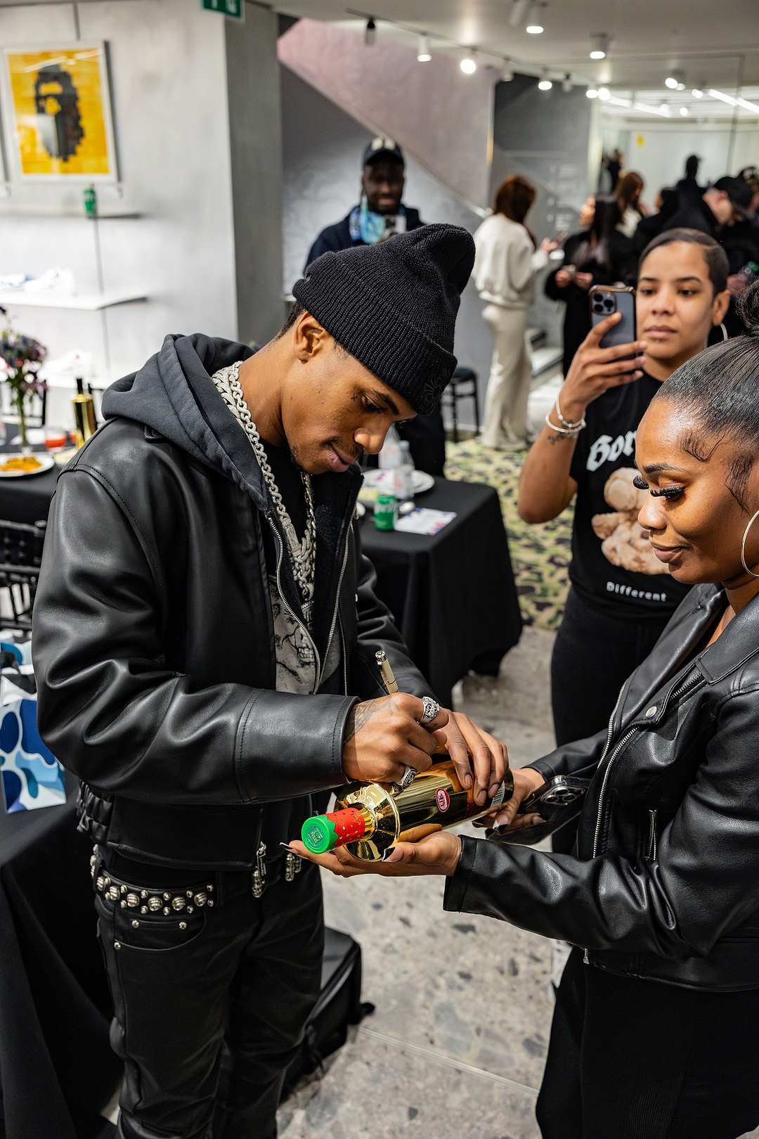 Man in black beanie and leather jacket signing a gold bottle held by a woman in a black leather jacket