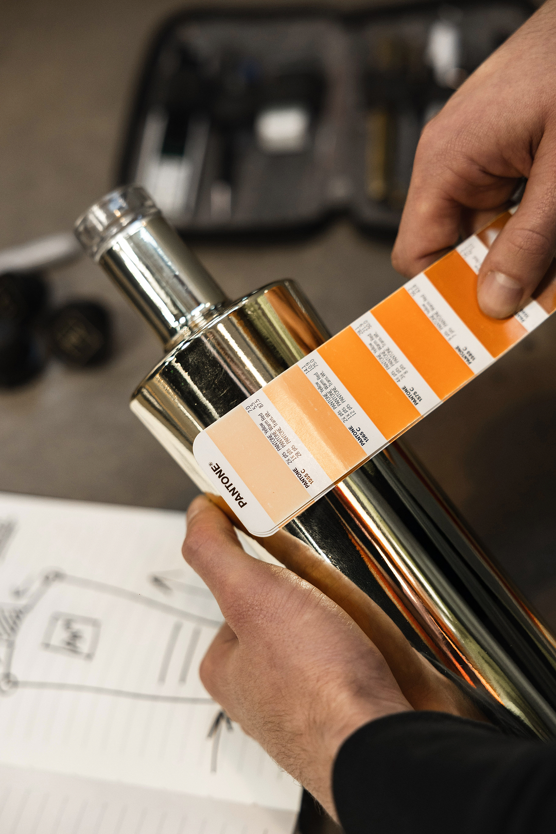Hands holding a Pantone color guide against a metallic bottle