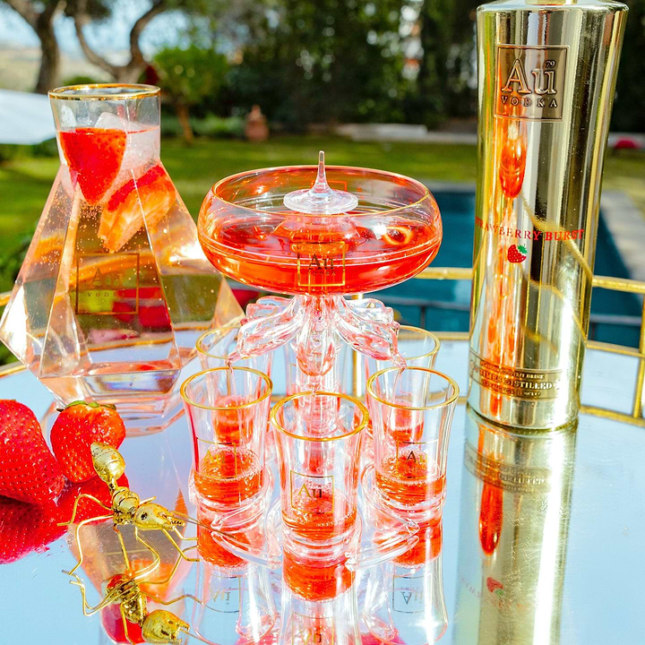 Au Vodka bottle next to a glass shot tower filled with red liquid on a mirrored tray with fresh strawberries