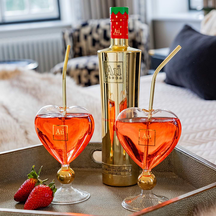 Au Vodka bottle and two heart shaped glasses with red cocktail and gold straws on a tray with fresh strawberries