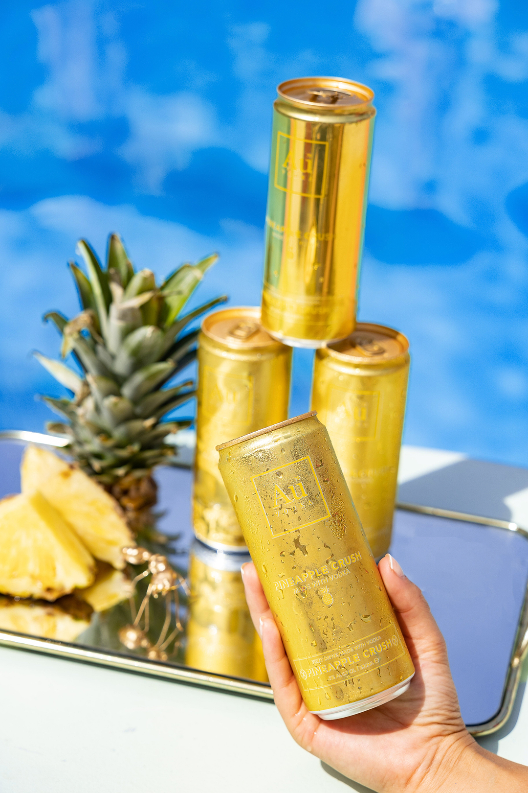 Hand holding a gold Au Vodka Pineapple Crush can with stacked cans and fresh pineapple on a mirrored tray