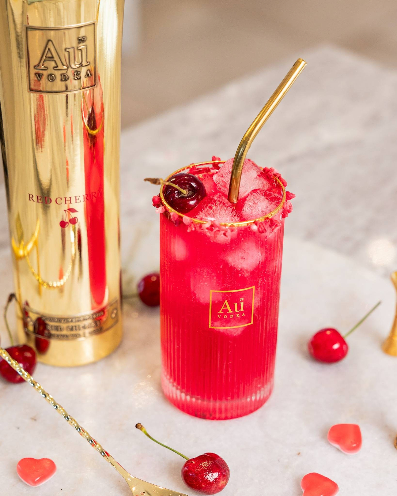 Au Vodka Red Cherry bottle next to a glass of red cocktail with ice, a gold straw, and fresh cherries