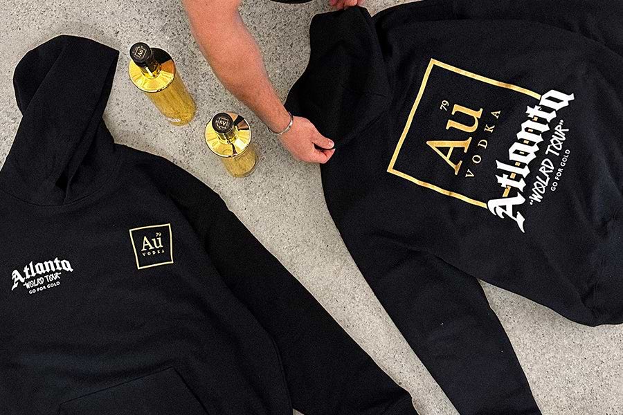 Two black hoodies with gold Au Vodka branding and two gold Au Vodka bottles on a concrete surface