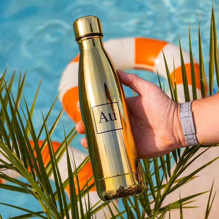 Hand holding a gold Au Vodka USA water bottle with a pool and palm leaves in the background