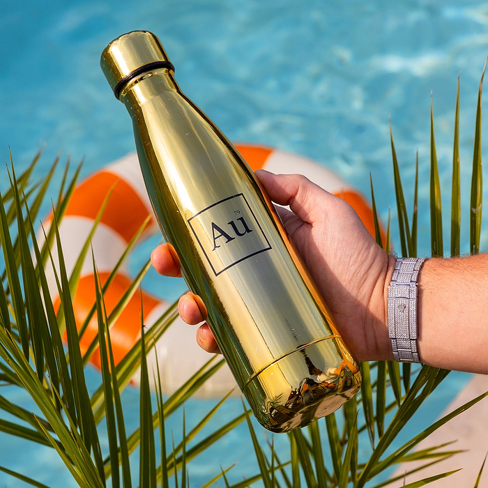 Hand holding a gold Au Vodka USA water bottle with a diamond watch, palm leaves, and a pool in the background
