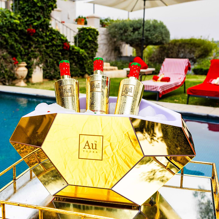 Three Au Vodka Red Cherry bottles in a gold diamond shaped ice bucket by a pool