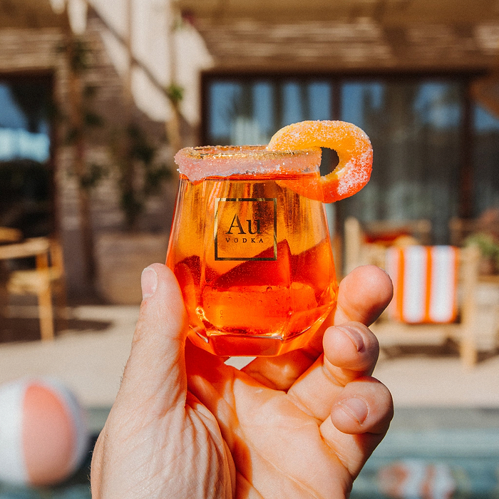 Hand holding an Au Vodka branded glassware filled with an orange cocktail and a sugar rimmed peach slice garnish