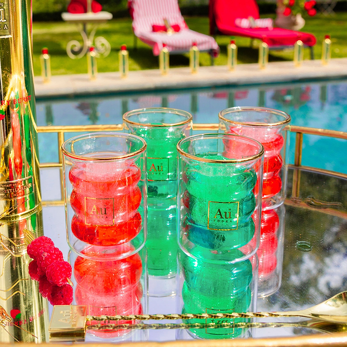 Four Au Vodka branded glassware tumblers filled with red and green drinks on a mirrored tray by a pool