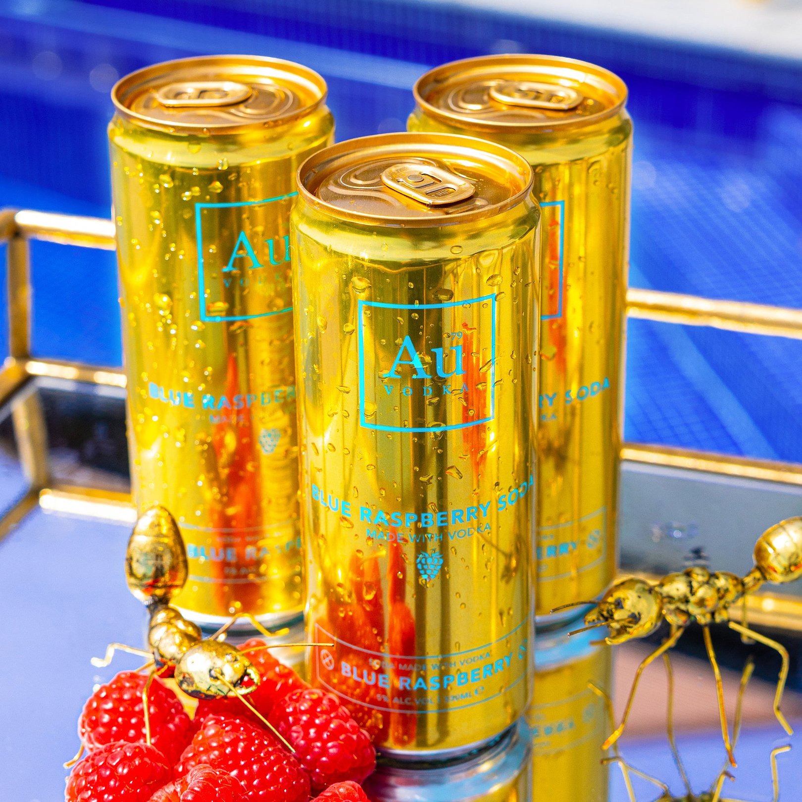 Three gold Au Vodka Blue Raspberry soda cans with fresh raspberries and gold ant ornaments