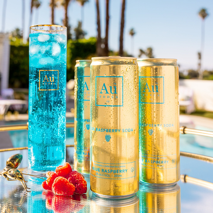 A glass of blue liquid next to three Au Vodka Blue Raspberry Soda cans and raspberries