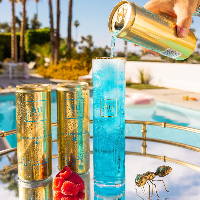 A hand pouring Au Vodka Blue Raspberry Soda into a glass next to two cans and raspberries