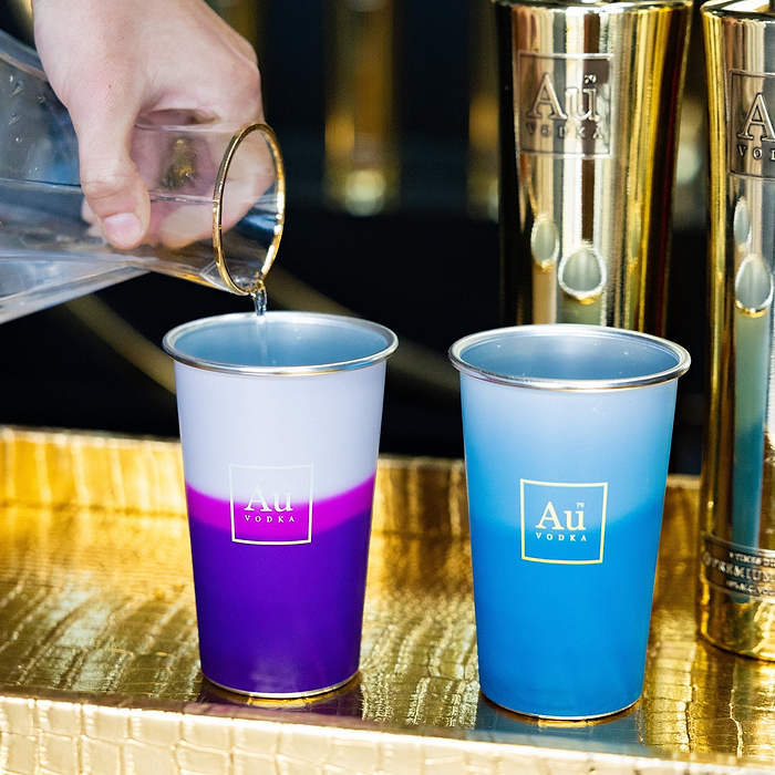 Hand pouring water into an Au Vodka USA purple and white cup, with a blue and white cup next to it
