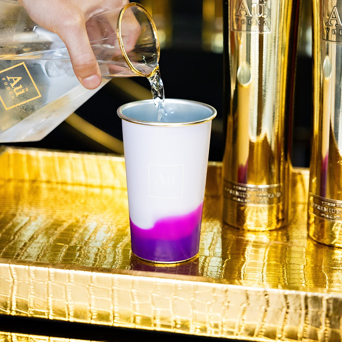 Hand pouring clear liquid from an Au Vodka bottle into a white and purple Au Vodka tumbler