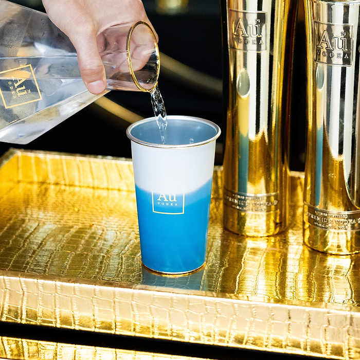 Hand pouring clear liquid from Au Vodka bottle into blue and white Au Vodka cup on gold tray
