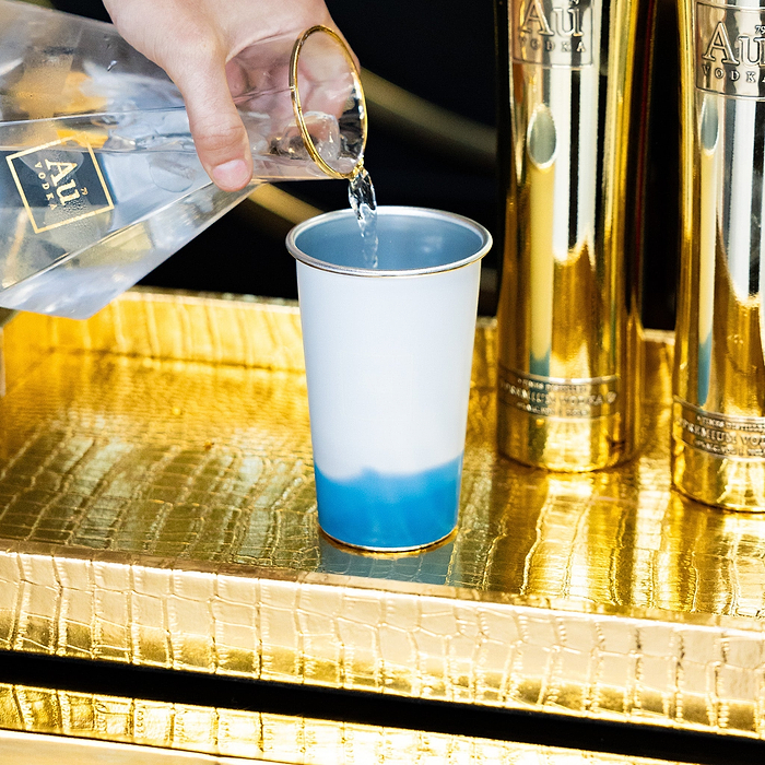 Hand pouring Au Vodka into a white cup with blue liquid, on a gold crocodile-textured tray