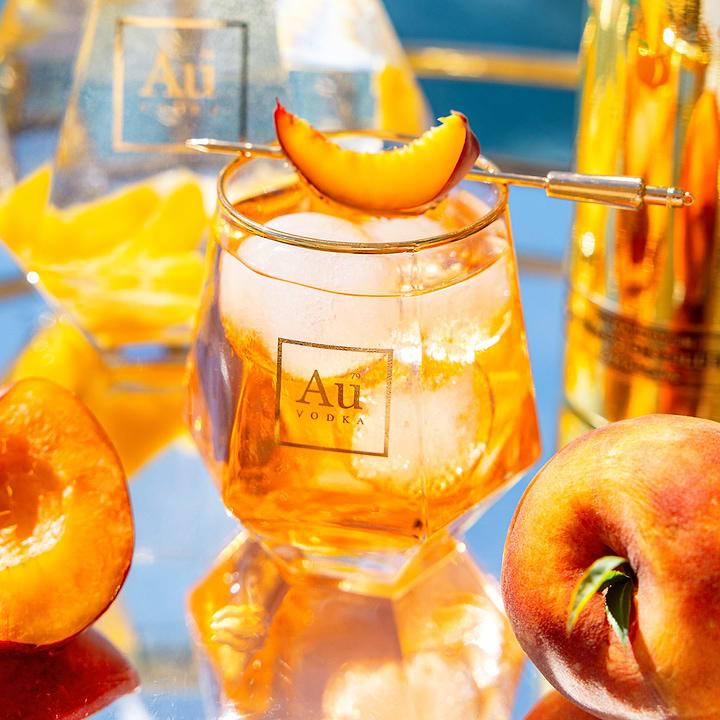 Au Vodka cocktail in a faceted glass with ice and a peach slice garnish on a reflective surface