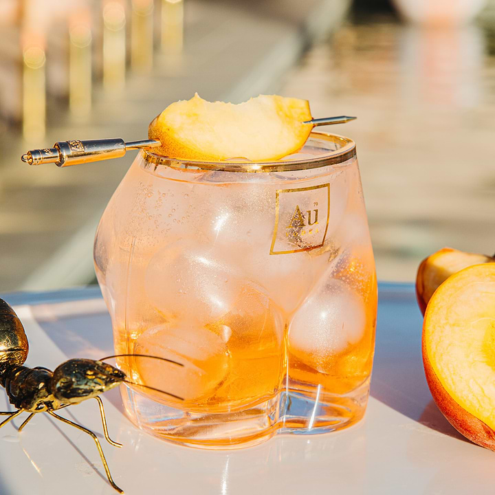 Au Vodka branded glassware filled with ice and a peach slice garnish on a gold cocktail pick
