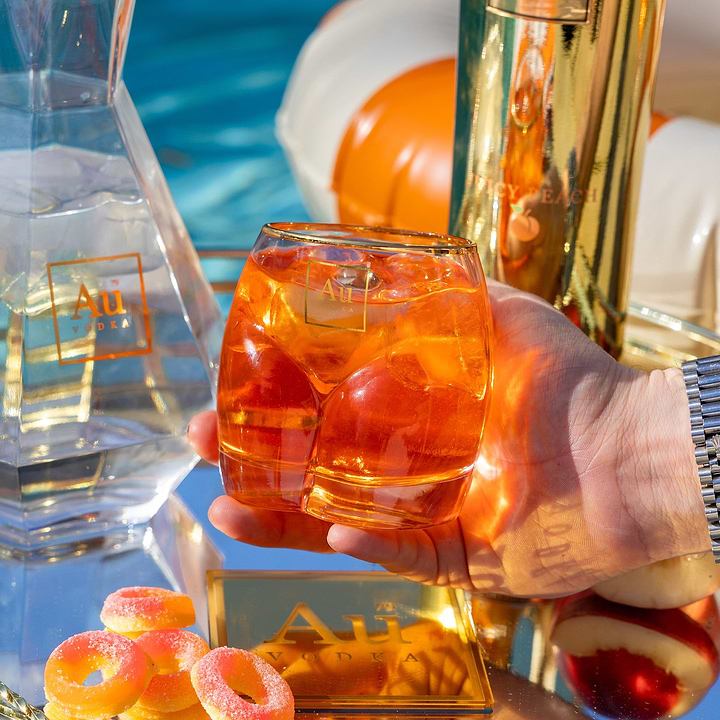 Au Vodka branded glassware filled with orange cocktail and ice held by hand