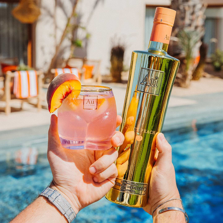 Au Vodka Juicy Peach bottle and branded glassware with peach garnish by a swimming pool