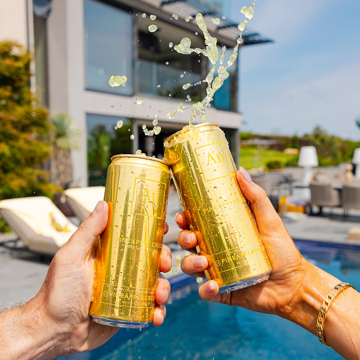 Two hands holding Au Vodka Pineapple Crush cans, liquid splashing from one can, by a pool